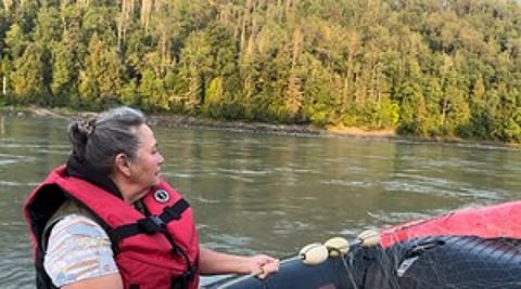 Hereditary Chief Darlene Simpson wears a life jacket while traveling by boat on a calm river lined with dense forest in the "traditional territory" of the Tsetsaut Skii km Lax Ha Nation.