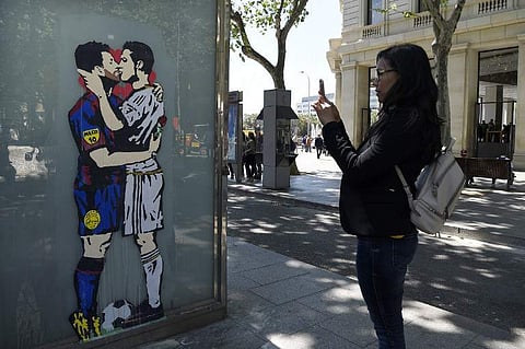 A woman takes a picture to the street art 'Love is blind' of Street artist Salva 'Tvboy' on April 21, 2017 in Barcelona, which shows Barcelona's Argentinian forward Lionel Messi and Real Madrid's Portuguese forward Cristiano Ronaldo kissing each other two