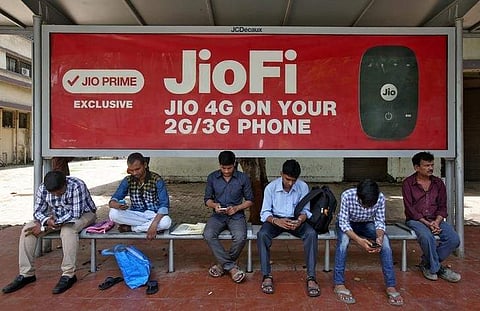 Commuters use their mobile phones as they wait at a bus stop with an advertisement of Reliance Industries' Jio telecoms unit, in Mumbai, on July 10. (Photo | Reuters)