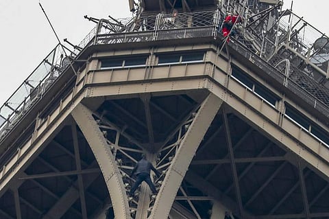 A man (C) climbs up to the top of the Eiffel Tower, in Paris, without any protection as a firefighter (R) looks down at him from the top. (Photo | AFP)