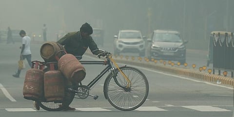A worker carrying LPG cylinders on a bicycle crosses a road, shrouded in a thick layer of smog, in New Delhi. (Photo | PTI)