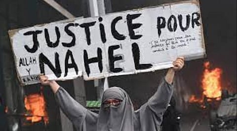 - An attendee holds a banner reading 'Justice for Nahel' as cars burn in the street at the end of a commemoration march for a teenage driver shot dead by a policeman. (Photo | AFP)