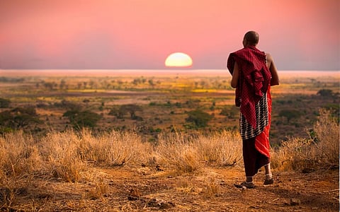 A Maasai man, wearing traditional blankets, overlooks Serengeti in Tanzania as the colourful sunset fills the sky. 