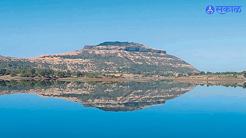 The reflection of the fort and surrounding hills in the water of Tringalwadi Dam at dawn.