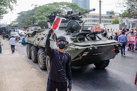 A protester holds a placard with the three-finger salute in front of a military tank in front of the Central Bank building during a demonstration. February 2021, Yangon, Myanmar. Photo: IMAGO / ZUMA Wire. 