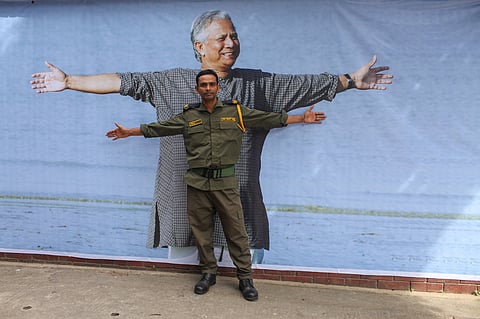 A man in a green security guard uniform stands with arms spread out in front of a poster of Muhammad Yunus in a grey checked kurta, also with arms spread out. 