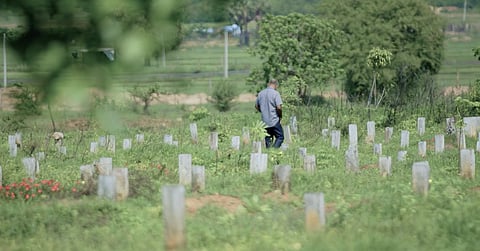 A still from the documentary ‘Oddamavadi’, where a mourner dressed in a grey shirt and black pants walks through an overgrown burial lot dotted with tombstones. 