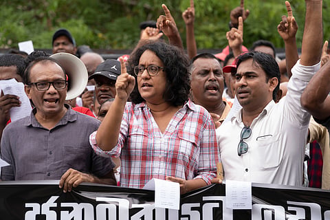 Harini Amarasuriya dressed in a red and white checked shirt standing behind a black banner with white lettering held by two male party members on either side during a protest march in June 2023.