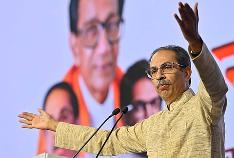 Maharashtra politician Uddhav Thackeray, wearing a beige kurta, stands in front of a mike with his arms spread out. In the background is a large banner with the image of his father Bal Thackeray wearing a saffron scarf.  