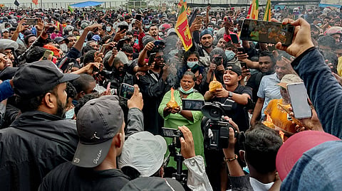 A large crowd of Aragalaya protesters gather around two individuals breaking coconuts. Several activists at the scene are holding phones and cameras to record the event. 