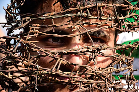 A close-up photograph of a man's face partially obscured by tangled and rusted barbed wire as he stare through the gaps. The background is slightly blurred, with hints of a green and white flag visible behind him. 