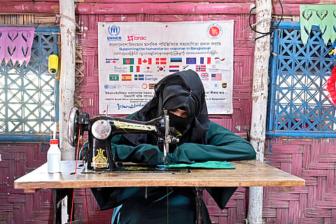 A Rohingya woman dressed in an abaya and hijab works at a sewing machine in a. refugee camp. A banner with logos of donor organisations hangs in the background.