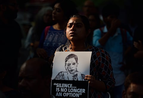 A woman stands in a crowd holding a black-and-white poster featuring a portrait of Gauri Lankesh and the words 'SILENCE... IS NO LONGER AN OPTION'. The background scene is a protest or vigil, with other people visible in the dimly lit background.