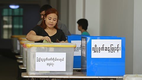 Woman casting a ballot in a box. A man can be seen behind her also casting a vote. The ballot box has writing on it in Burmese. 