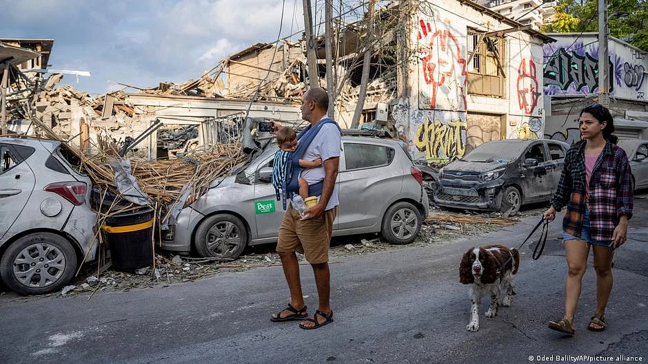 A family with baby in a carrier and a pet dog walk past a Tel Aviv building reduced to rubble by a rocket, after Hamas launched a multi-front attack on Israel starting 7 October (photo: Oded Balilty/AP/picture alliance via DW)