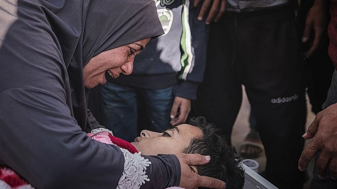 A mother in black abaya and hijab cries over her dead child in Gaza; beside her, just outside the frame, her younger child stands crying too (photo courtesy @azaizamotaz9/X)