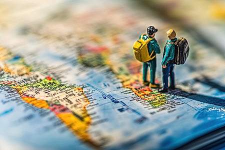 Close-up captures two tiny traveler figurines with vibrant backpacks standing atop a world map, seemingly planning their journey across continents