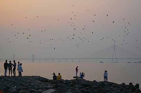 People sit along the shoreline at Dadar Beach as the Bandra-Worli Sea Link remains shrouded in smog, in Mumbai, Thursday, Nov. 27, 2025. 