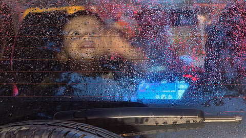 A child gazes through a rain-speckled rear window as evening showers and city lights create a dreamy monsoon scene at Thiruvananthapuram