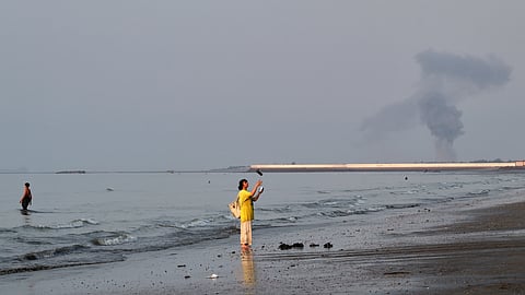 A woman stands at the water’s edge along the Strait of Hormuz as a plume of smoke rises in the background following an explosion off Bandar Abbas, Iran on July 13