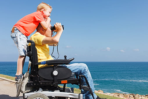 A father in a motorized wheelchair carries a smiling boy on his shoulders.
