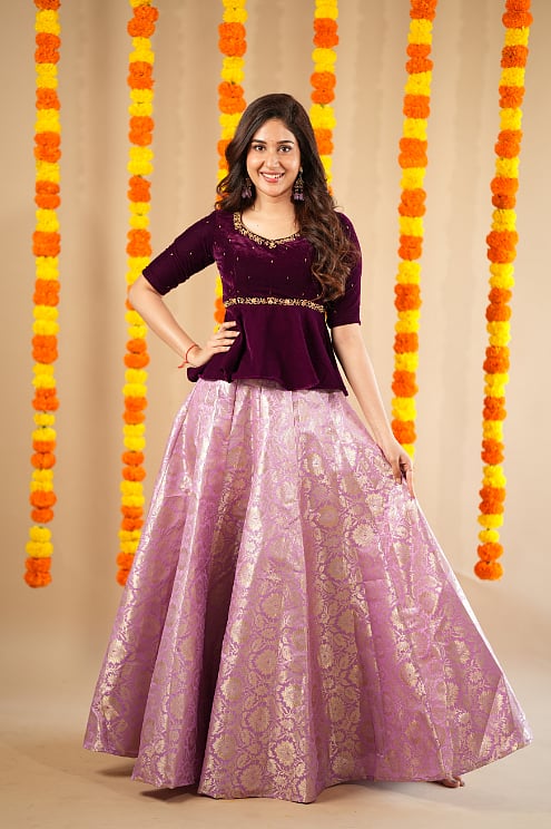 A woman in purple traditional dress stands in front of a floral background.
