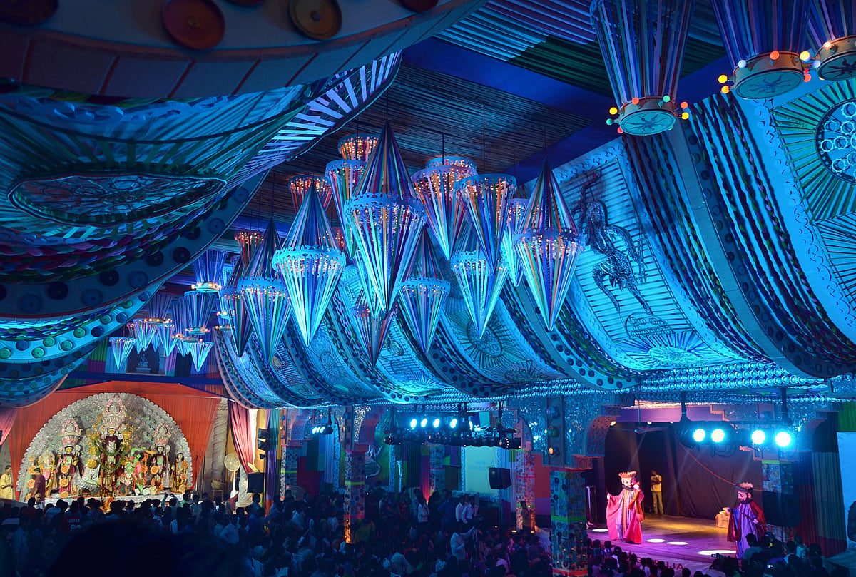 An ornate pandal with a blue draped ceiling and a stage with performers and an idol.