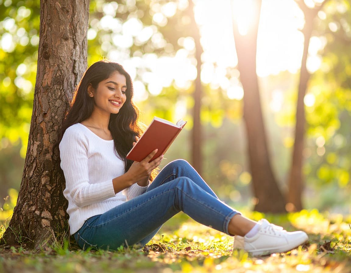 Woman sitting under a tree reading a book and smiling in a sunny outdoor setting.