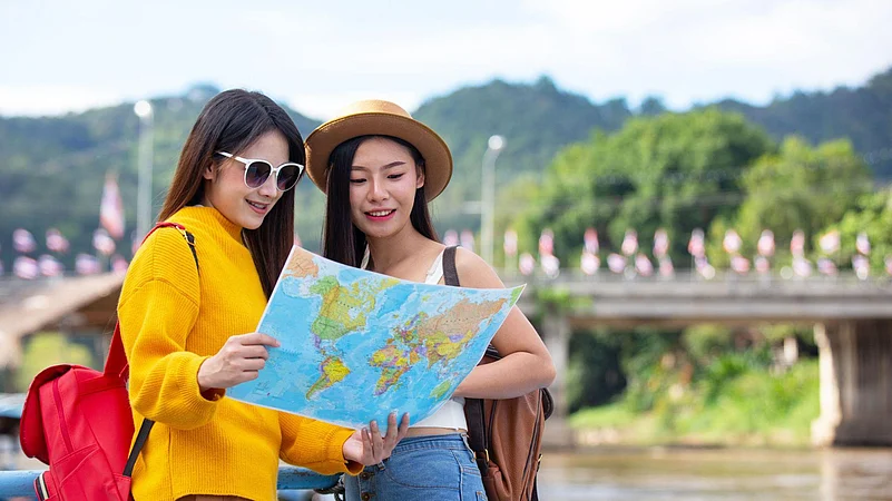 Two women travelers with backpacks looking at a world map near a river bridge.
