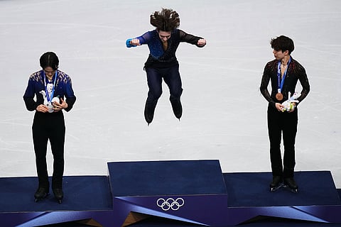 Gold medalist Mikhail Shaidorov of Kazakhstan jumps up to the podium to receive his medal with silver medalist Yuma Kagiyama of Japan, left, and bronze medalist Shun Sato of Japan, right, after competing in the men's free skate program in figure skating at the 2026 Winter Olympics, in Milan, Italy.
