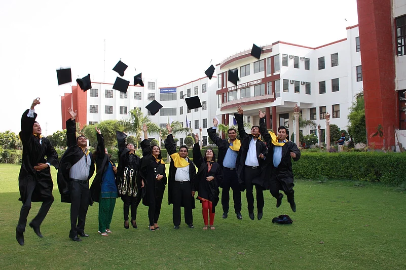 Graduates in black robes tossing their caps in the air in front of a white campus building.
