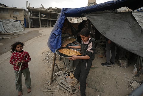 A barefoot Palestinian girl looks at another girl carrying a tray of baked traditional Eid cookies outside a partially destroyed home in Gaza City, Wednesday, March 18, 2026. 