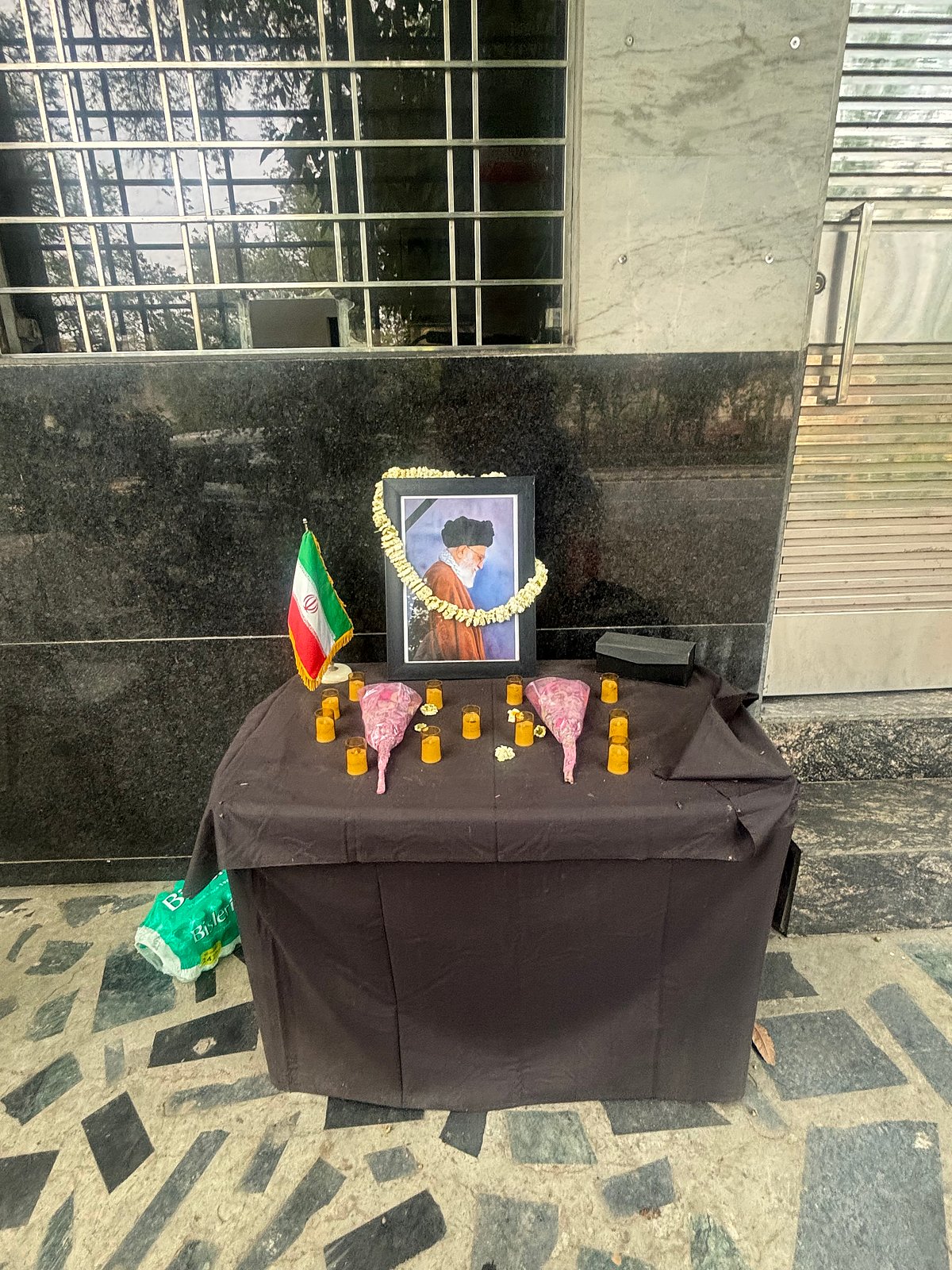 Fozia Yasin : Outside the gates, a small table has been set up. On it rests a photograph of Ayatollah Ali Khamenei, framed by flowers. 