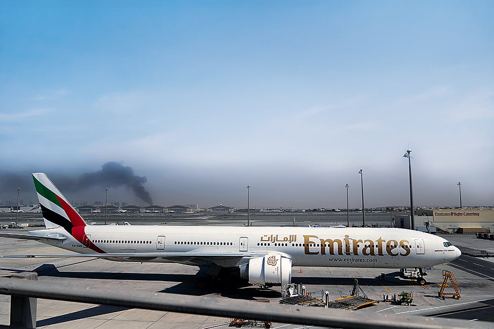 Enveloped by Darkness: A plume of smoke caused by an Iranian strike is seen in the background of an Emirates plane parked at the Dubai International Airport on March 1, 2026 - | Photo: AP
