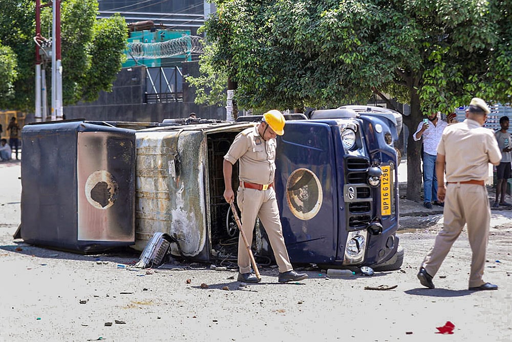 | Photo: PTI : Police personnel inspect the wreckage of a vehicle during a protest by factory workers demanding a hike in wages, in Noida, Gautam Buddh Nagar district, Uttar Pradesh. The protest carried incidents of arson, vandalism and stone-pelting reported from Phase-2 and Sector 60 areas, police said. 