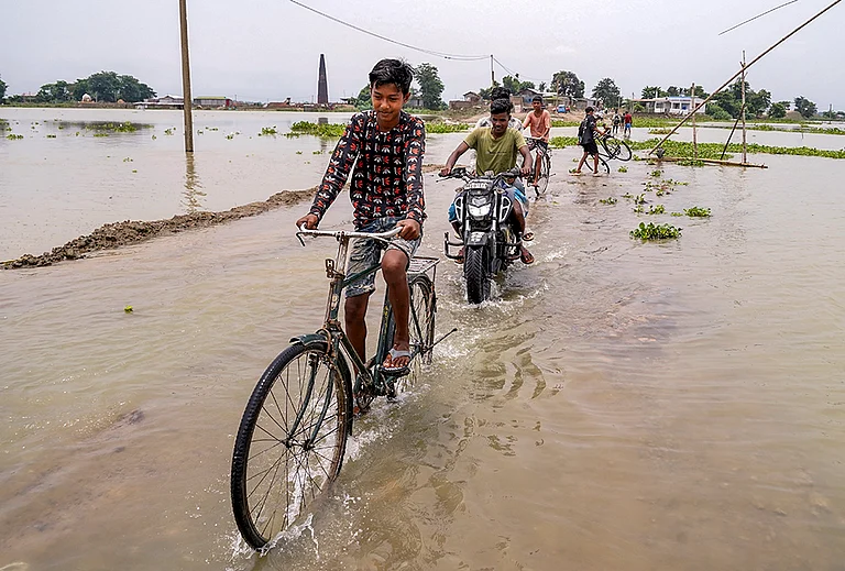 People wade through a waterlogged road at Morisutipam village in Morigaon district, Assam. - | Photo: PTI