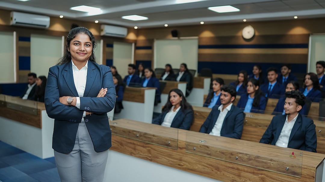 A smiling woman in a grey blazer stands in a university lecture hall filled with seated students