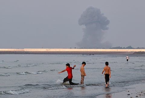 Three boys play in the shallow waters of the Strait of Hormuz, as a plume of smoke rises from an explosion in the background, off Bandar Abbas, Iran.