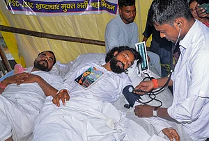 | Photo: PTI : A medical worker checks the blood pressure of student leader Devendra Nath Mahto as he lies with a copy of the Constitution of India on his chest during an indefinite hunger strike at Jaipal Singh Munda Stadium, in Ranchi, Wednesday, Aug. 5, 2026. Mahto and fellow protesters are demanding a probe into alleged recruitment exam irregularities conducted by the JPSC and JSSC.