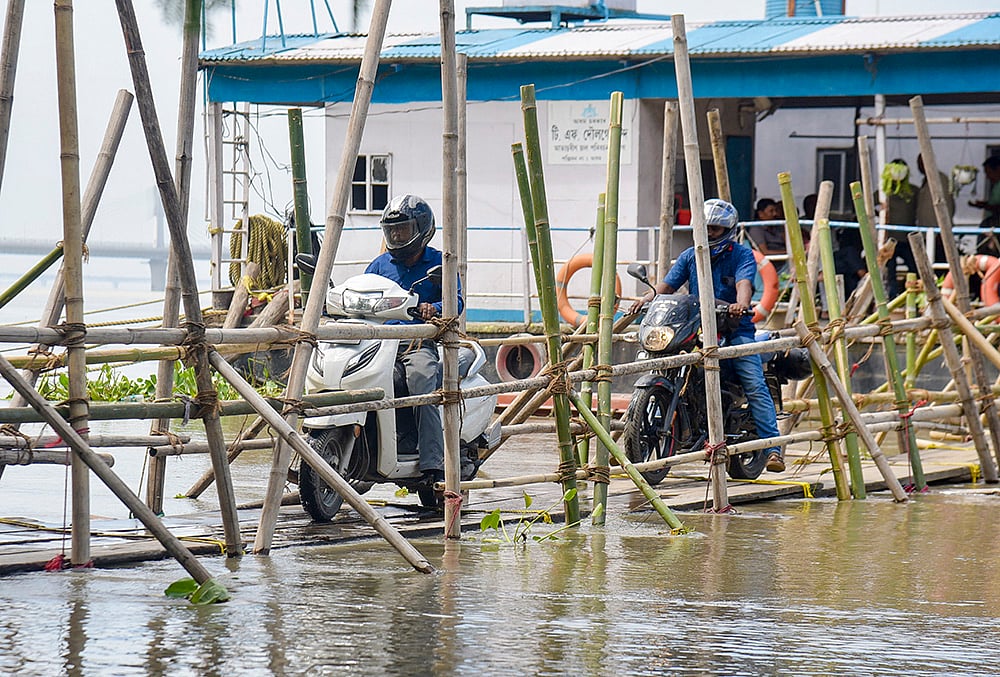 Commuters ride two-wheelers across a makeshift bamboo bridge to reach a ferry terminal over the Brahmaputra River, in Guwahati. - | Photo: PTI