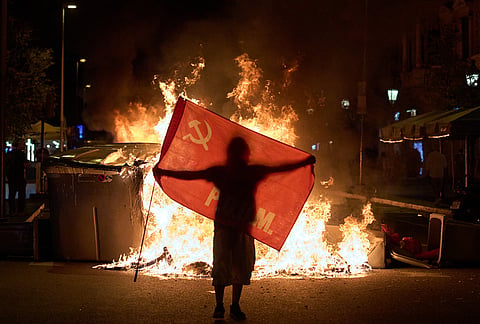 A protesters holds a flag next to trash cans on fire during clashes with police following a demonstration over Spain's housing crisis, hours after Prime Minister Pedro Sánchez announced an early general election, in Barcelona, Spain, Monday, Oct. 5, 2026. 

