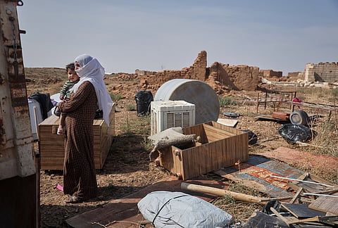 A woman holds a young child amid household belongings and destroyed buildings after returning to Al-Arisha, in Syria's Ras al-Ayn countryside, Tuesday, Oct. 13, 2026, seven years after a Turkish military offensive alongside allied Syrian factions forced residents to flee.