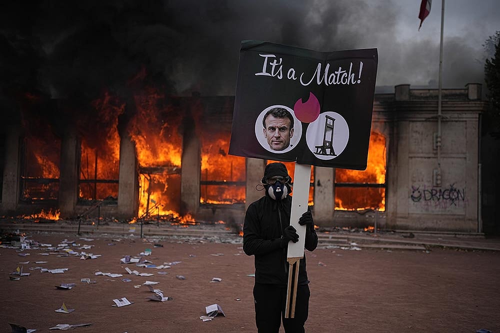 A protester holds a board with a photo of French president Emmanuel Macron in front of a burning tourism office during a demonstration against rising living costs and in solidarity with students’ grievances in Lyon, France, Saturday, Oct. 17, 2026. - | Photo: AP/Laurent Cipriani