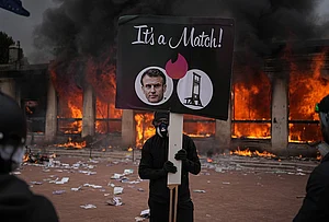 | Photo: AP/Laurent Cipriani : A protester holds a placard with a photo of French president Emmanuel Macron and a guillotine in front of a burning tourism office during a demonstration against rising living costs and in solidarity with students' grievances in Lyon, France, Saturday, Oct. 17, 2026.