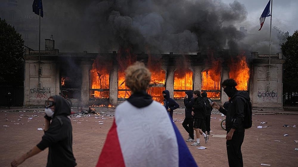 | Photo: AP/Laurent Cipriani : A protester wraps a French flag around them in front of the burning tourism office during a demonstration against rising living costs and in solidarity with students’ grievances in Lyon, France, Saturday, Oct. 17, 2026.