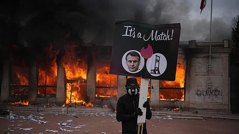 A protester holds a board with a photo of French president Emmanuel Macron in front of a burning tourism office during a demonstration against rising living costs and in solidarity with students’ grievances in Lyon, France - | Photo: AP/Laurent Cipriani
