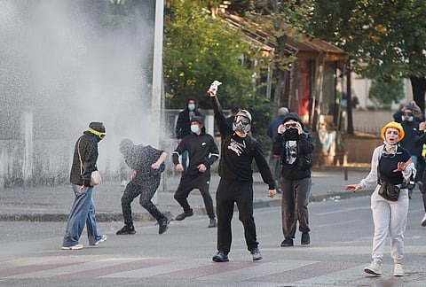A protester in a gas mask gestures as a police water cannon sprays nearby during an anti-government protest in Tirana, Albania, Tuesday, Oct. 20, 2026. 