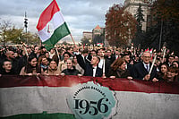 In Photos: Hungary Marks 70 Years Since 1956 Revolution Against Soviet Rule | Photo: Zoltan Kocsis/MTI via AP : Hungarian Prime Minister Peter Magyar, center, waves a Hungarian flag with a hole cut in its middle, which symbolises the Hungarian revolution in 1956, during the traditional torchlight memorial march of the 70th anniversary of the outbreak of the Hungarian Revolution and Freedom Fight of 1956 against Soviet oppression in front of the Budapest University of Technology and Economics in Budapest, Hungary, Thursday, Oct. 22, 2026.