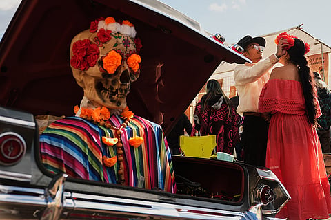 Marc Bautista helps Cassandra Harrisson with her outfit as they stand next to their 1963 Chevy Impala during a Día de Muertos celebration at Pike Place Market, Friday, Oct. 23, 2026, in Seattle.