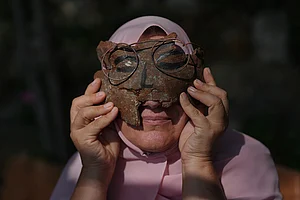 | Photo: AP/Hassan Ammar : Visual artist Lina Jouni holds an artwork depicting a face wearing glasses, created from shrapnel collected from Israeli military munitions, over her face at her home in the village of Roumin, southern Lebanon, Sunday, Oct. 25, 2026.